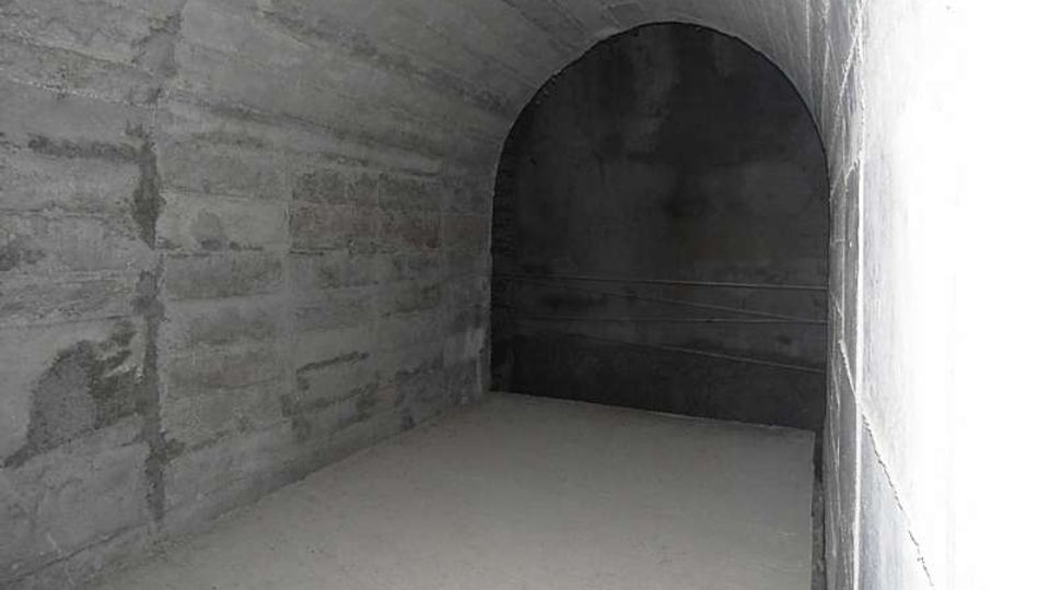 Interno di un tunnel sotterraneo in cemento grezzo, con pareti e soffitto curvi e pavimento spoglio. In fondo si vede un’apertura ad arco che conduce a una zona più buia.