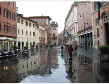 Ampia piazza urbana con pavimentazione in pietra bagnata dalla pioggia, circondata da edifici storici dai colori pastello e facciate con finestre regolari. Ai lati si vedono tavolini di un bar con ombrelloni, portici con colonne, lampioni e grandi fioriere.