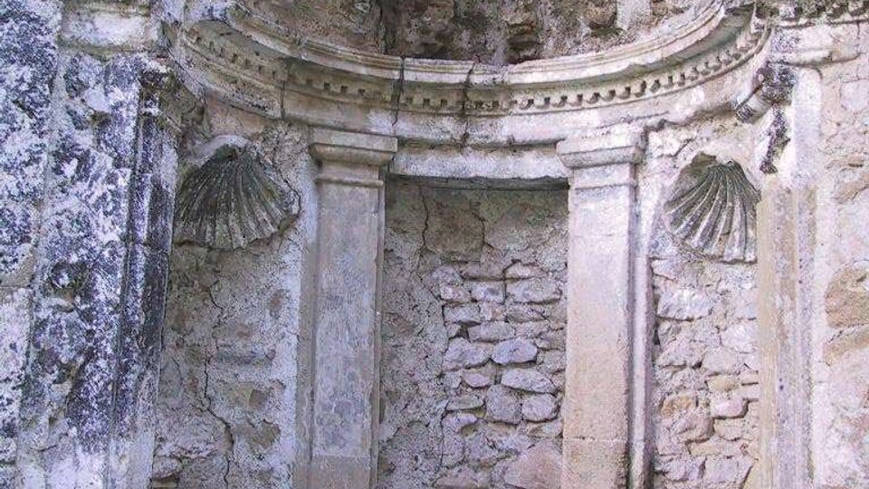 Nicchia architettonica in pietra in un muro antico e deteriorato, con due pilastri, cornice modanata e conchiglie decorative laterali. Sopra la nicchia si vede una semicupola intonacata e screpolata, con vegetazione alla base.