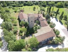 Veduta aerea di un complesso rurale in pietra con tetti in coppi rossi e una torre centrale, circondato da alberi e giardini. Intorno si vedono prati verdi, filari di cipressi e un’area di parcheggio asfaltata sul lato destro.