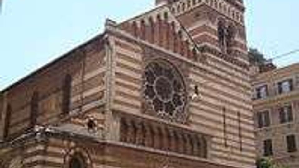 Facciata di una chiesa romanico-gotica in pietra a fasce orizzontali bianche e scure, con grande rosone centrale e portale ad arco. Sul lato si innalza un campanile quadrato a più livelli, affacciato su una piazza urbana con edifici circostanti.