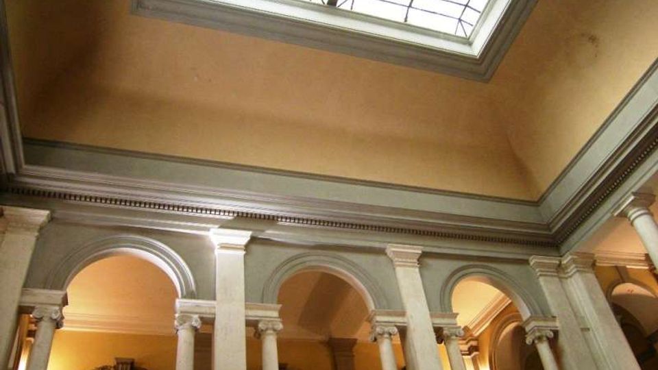 Interno di un edificio storico con cortile o atrio a due livelli, caratterizzato da una serie di arcate e colonne in pietra. In alto si vede un grande lucernario rettangolare con struttura in vetro e cornice decorata che illumina lo spazio.