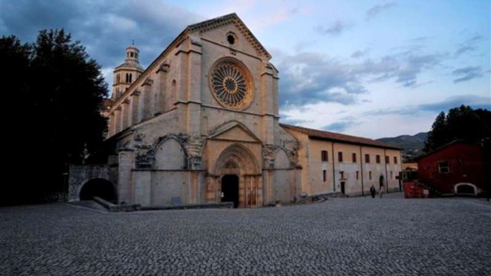 Facciata di una grande chiesa in pietra con rosone centrale e portale ad arco, affacciata su un’ampia piazza lastricata. A lato si estende un lungo edificio adiacente, con alberi e colline sullo sfondo sotto un cielo nuvoloso.