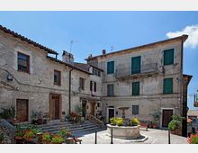 Piccola piazza in un borgo con case in pietra e intonaco, finestre con persiane verdi e balcone in ferro. Al centro c’è una fontana circolare in pietra, con gradini, panchine e fioriere, sotto un cielo sereno.