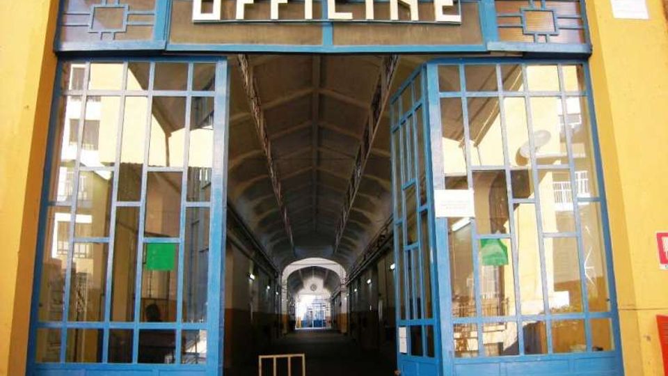 Ingresso di un edificio industriale con grandi porte vetrate blu e insegna “OFFICINE” sopra l’architrave. All’interno si vede un lungo corridoio coperto con soffitto alto ad arco e luce in fondo al passaggio.
