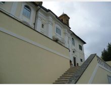 Facciata di un edificio storico chiaro con decorazioni in stucco e finestre ad arco, vista dal basso lungo una scalinata esterna con parapetto. Sul tetto si intravede una piccola torretta e sullo sfondo c’è cielo coperto con vegetazione laterale.