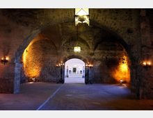 Ampia sala sotterranea in pietra con soffitto a volte e grandi archi, illuminata da lanterne sospese e applique a parete con luce calda. In fondo, un arco centrale conduce a un cortile esterno più luminoso.