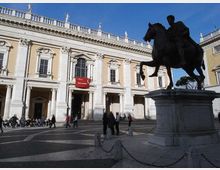 Piazza con grande statua equestre in bronzo su alto basamento di pietra, in primo piano. Sullo sfondo si vede un palazzo storico in stile classico con facciata chiara, portico ad archi, finestre ornate e balaustra con statue sul tetto.