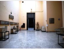 Sala museale con pavimento in pietra chiara e alte pareti color crema, con lapidi e reperti archeologici esposti su supporti e lungo le pareti. Al centro si apre un grande portale scuro che conduce a un ambiente interno più buio.