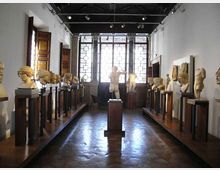 Interno di una galleria museale con pavimento in legno e file di busti e sculture su piedistalli lungo le pareti. In fondo, grandi finestre con vetri decorati illuminano la sala e una statua centrale su basamento è posizionata nell’asse del corridoio.