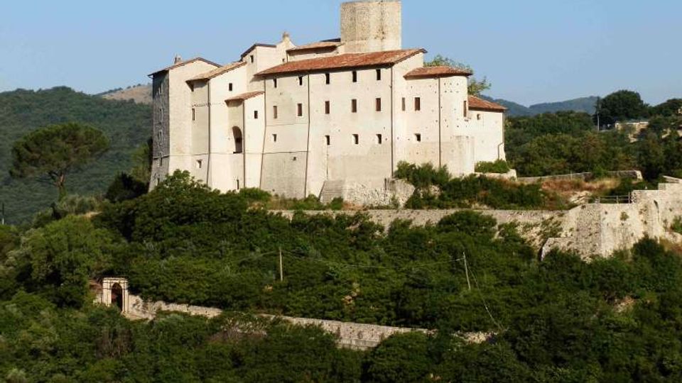 Grande castello in pietra chiara su una collina, con mura massicce, poche finestre e un torrione cilindrico centrale, coperto da tetti in tegole rosse. Intorno si vedono boschi e vegetazione fitta, con un muro di cinta e un ingresso ad arco in basso.