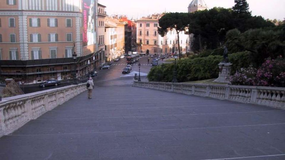 Ampia scalinata in pietra con balaustre che scende verso una piazza urbana, circondata da palazzi storici e una strada con auto. Sullo sfondo si vedono alberi di un parco e una cupola in lontananza.