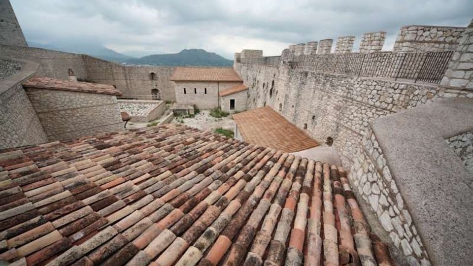 Veduta dall’alto dell’interno di una fortezza in pietra, con tetti in coppi di terracotta e mura merlate lungo il perimetro. Sullo sfondo si vedono montagne e cielo nuvoloso.