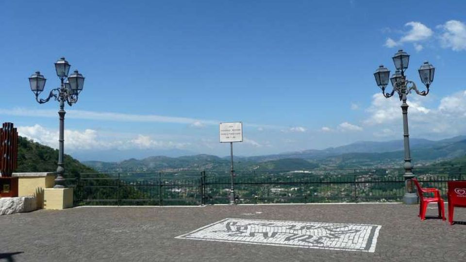 Piazzale panoramico pavimentato in pietra con ringhiera affacciata su una valle e colline lontane. Due lampioni in ferro battuto e un cartello centrale incorniciano la vista sotto un cielo azzurro.