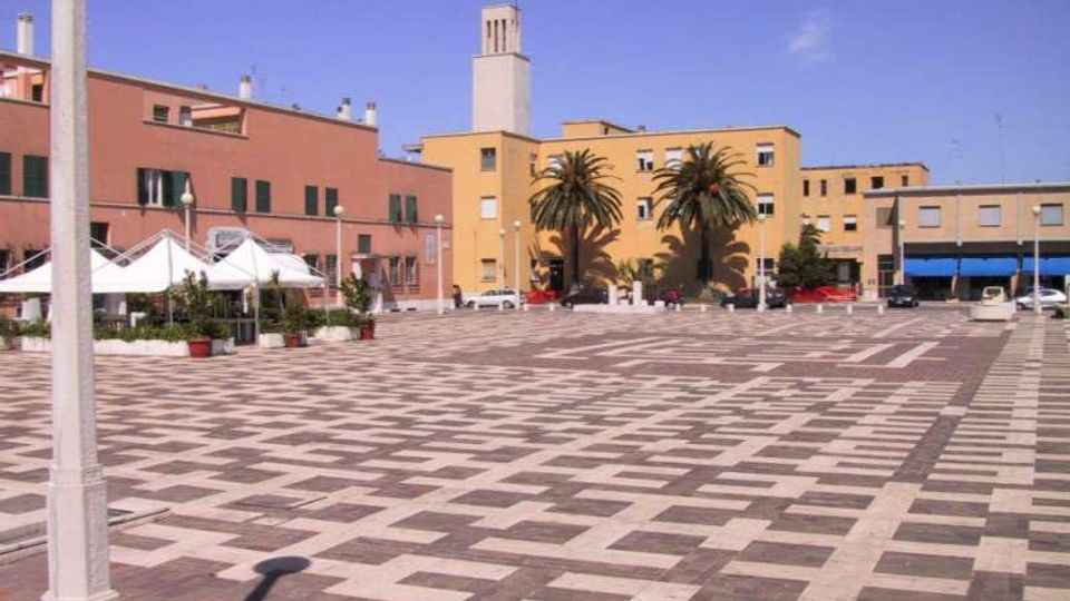 Ampia piazza urbana pavimentata con motivi geometrici in pietra chiara e scura, circondata da edifici bassi color ocra e rosso. Sullo sfondo si vedono una torre campanaria, alcune palme e strutture con tende bianche lungo il lato sinistro.