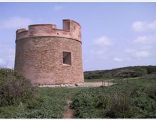 Torre cilindrica in mattoni con sommità merlata e un’apertura rettangolare, situata in una zona di campagna con vegetazione bassa e sentiero sterrato. Sullo sfondo si vedono colline verdi sotto un cielo parzialmente nuvoloso.