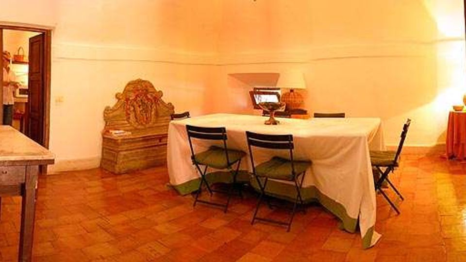 Interno di una sala con pavimento in cotto e soffitto a volta, con un lungo tavolo coperto da tovaglia bianca e sedie pieghevoli. Nella stanza si vedono una credenza in legno intagliato, un tavolo laterale e una porta in legno aperta verso un ambiente adiacente.