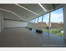 Ampio interno moderno e vuoto, con pavimento in cemento e soffitto con lunghe luci lineari, affacciato su una parete vetrata inclinata a tutta altezza. Attraverso le finestre si vedono edifici residenziali e alberi all’esterno.