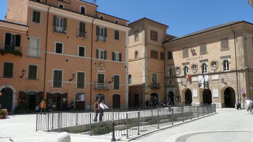 Piazza urbana lastricata con area centrale recintata da ringhiere metalliche, circondata da edifici storici a più piani con facciate color ocra e finestre con persiane. Sul lato destro si vede un palazzo con portici ad archi e stemmi sulla facciata, con bandiere esposte.
