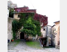 Cortile in ciottolato con edificio in pietra e intonaco, parzialmente coperto da edera verde e vite rossa, con un grande arco d’ingresso e finestre piccole. A destra una rampa con ringhiera conduce a un passaggio sottostante tra muri in pietra e lampioni.