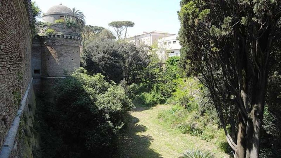 Veduta di un fossato verde con prato e vegetazione fitta ai piedi di alte mura in mattoni, con una torre cilindrica merlata sulla sinistra. Sullo sfondo si vedono alberi alti e alcuni edifici oltre il perimetro murario.