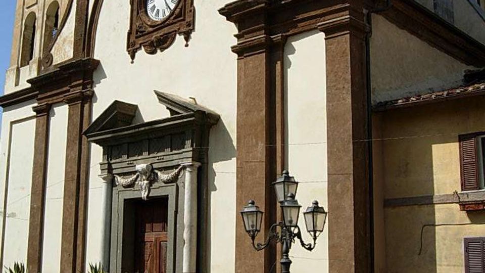 Facciata di una chiesa storica con intonaco chiaro e cornici in pietra scura, grande orologio centrale e portale rialzato con scalinata e ringhiera. Davanti si vedono un lampione a tre lanterne, vasi con piante e case adiacenti sotto un cielo sereno.