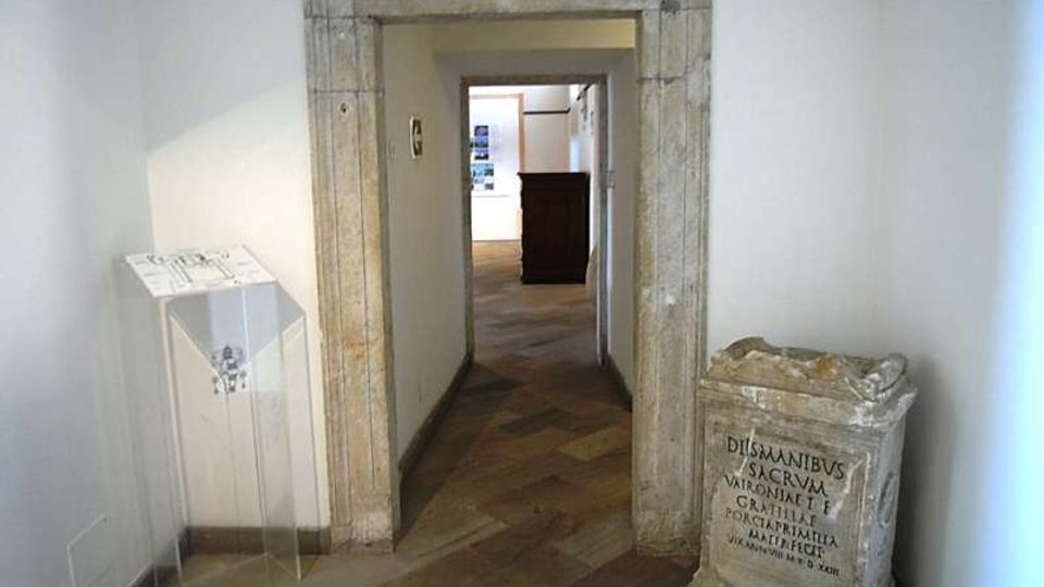 Corridoio interno con pareti bianche e pavimento in pietra, attraversato da un grande portale rettangolare in pietra che conduce a un’altra sala. Ai lati si vedono un leggio trasparente e un basamento in pietra con iscrizione, in un ambiente dall’aspetto museale.