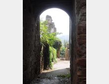 Passaggio in pietra sotto un arco, con pavimentazione acciottolata e muri in mattoni. Oltre l’arco si vede un sentiero con vegetazione rampicante e una torre in rovina sullo sfondo, con colline lontane.