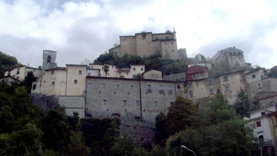 Borgo collinare con case in pietra addossate al pendio e un grande castello o fortezza sulla sommità. In primo piano si vede vegetazione fitta, mentre sullo sfondo il cielo è nuvoloso.