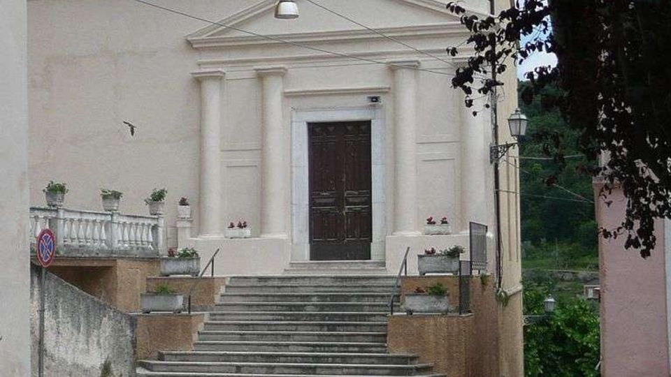 Facciata di una chiesa su una piccola strada in salita, con scalinata centrale in pietra che conduce a un portale scuro incorniciato da colonne e frontone. Ai lati ci sono muretti con fioriere e un parapetto, con cavi aerei e alberi sullo sfondo.