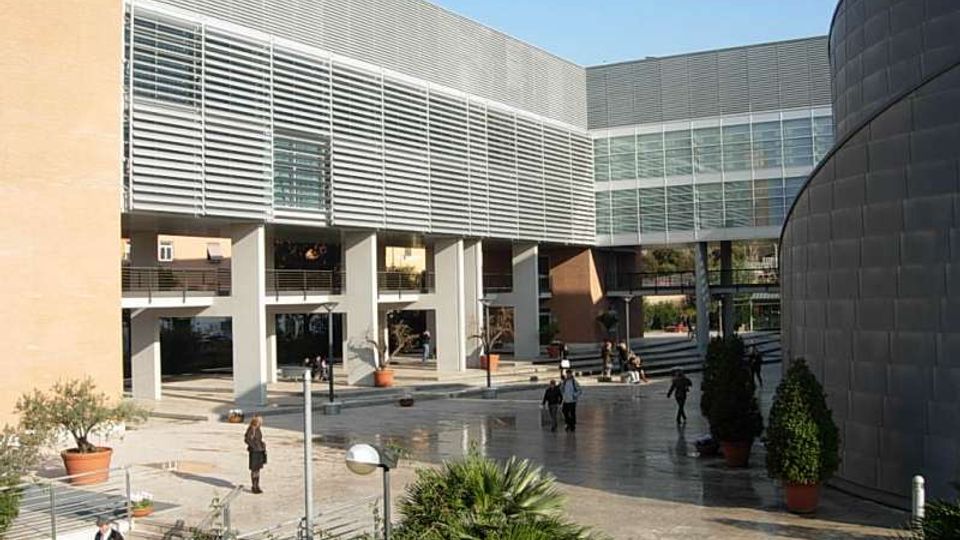 Cortile esterno di un complesso moderno con edifici rivestiti in metallo e grandi facciate vetrate, collegati da un passaggio sopraelevato. La piazza pavimentata include gradinate, lampioni e grandi vasi con piante e arbusti.