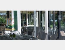 Terrazza coperta di un ristorante con tavoli apparecchiati e sedie intrecciate, affacciata su grandi vetrate con porte scorrevoli. Attraverso i vetri si vede un’area verde esterna con alberi e prato.