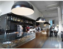 Interno di un bar moderno con lungo bancone in legno e vetrina espositiva, retrobanco nero con scaffali di bottiglie e attrezzature. Grandi lampade a sospensione a cupola illuminano lo spazio con pareti bianche e soffitto alto.