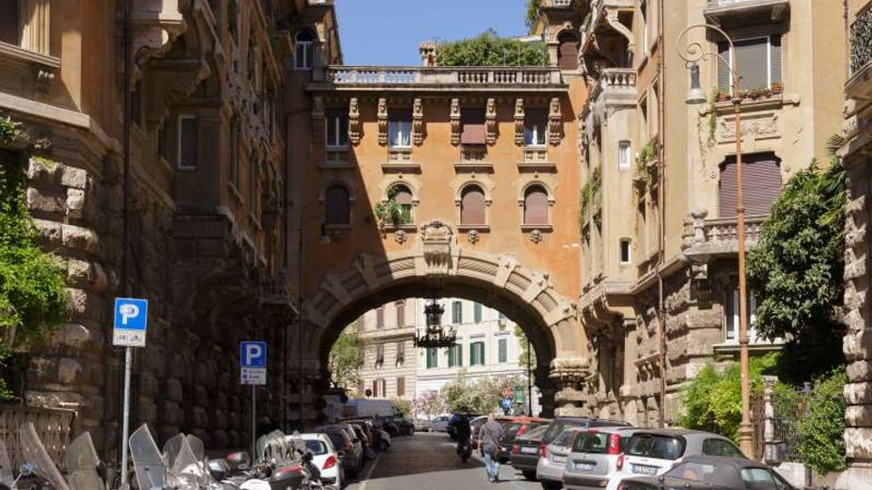 Strada urbana fiancheggiata da alti palazzi storici con facciate in pietra, balconi e finestre ad arco, con auto e moto parcheggiate lungo i lati. In fondo si vede un grande arco in muratura che collega gli edifici e incornicia la vista di un altro tratto di città oltre il passaggio.