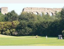 Campo da golf con ampio prato verde in primo piano e alberi fitti sullo sfondo. Dietro la vegetazione si intravedono i resti di un grande acquedotto in pietra con arcate.