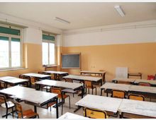 Aula scolastica con file di banchi e sedie rivolti verso una lavagna, pareti bicolori e illuminazione a neon sul soffitto. Grandi finestre laterali con tapparelle verdi e un tavolo dell’insegnante lungo la parete di fondo.