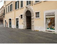 Facciata di un edificio storico color ocra lungo una strada lastricata, con grande portale ad arco in pietra bugnata e finestre con persiane. A destra si vede una vetrina di negozio con insegna “CORREDI” e scritta “SALDI”.