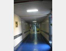 Lungo corridoio interno di un ospedale con pavimento blu lucido, pareti chiare con fascia grigia e illuminazione a plafoniere rettangolari sul soffitto. In fondo si vedono carrelli di servizio e porte lungo i lati del corridoio.