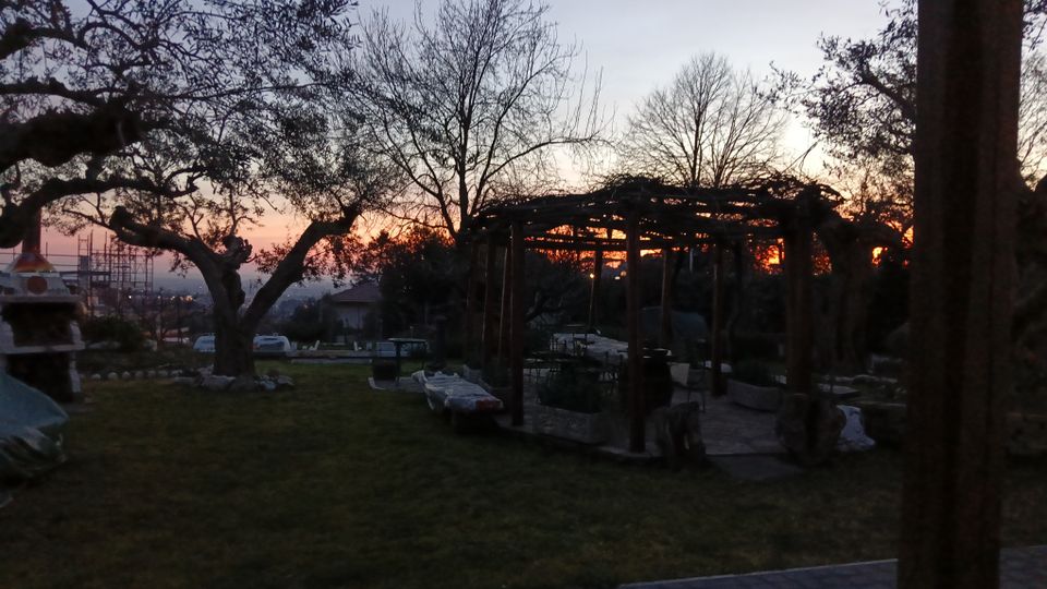 Giardino in collina al tramonto con prato, alberi spogli e un pergolato in legno con sedute e tavoli in pietra. Sullo sfondo si vedono la valle e alcune strutture lontane sotto un cielo sfumato arancione e blu.