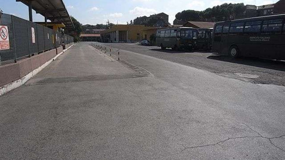 Ampio piazzale asfaltato di un deposito autobus, con pensilina e recinzione sul lato sinistro e vari autobus parcheggiati lungo il lato destro. Sullo sfondo si vedono edifici bassi e una collina alberata sotto un cielo sereno.