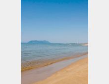 Spiaggia sabbiosa con battigia in primo piano e mare calmo e trasparente che si estende verso l’orizzonte. In lontananza si vede una costa bassa e una sagoma montuosa sotto un cielo azzurro limpido.