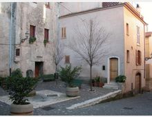 Piccola piazzetta lastricata in un borgo, con panchine verdi, vasi con arbusti e due alberi spogli. Intorno si affacciano case in muratura con intonaco chiaro, finestre con persiane e una porta ad arco sopra alcuni gradini.