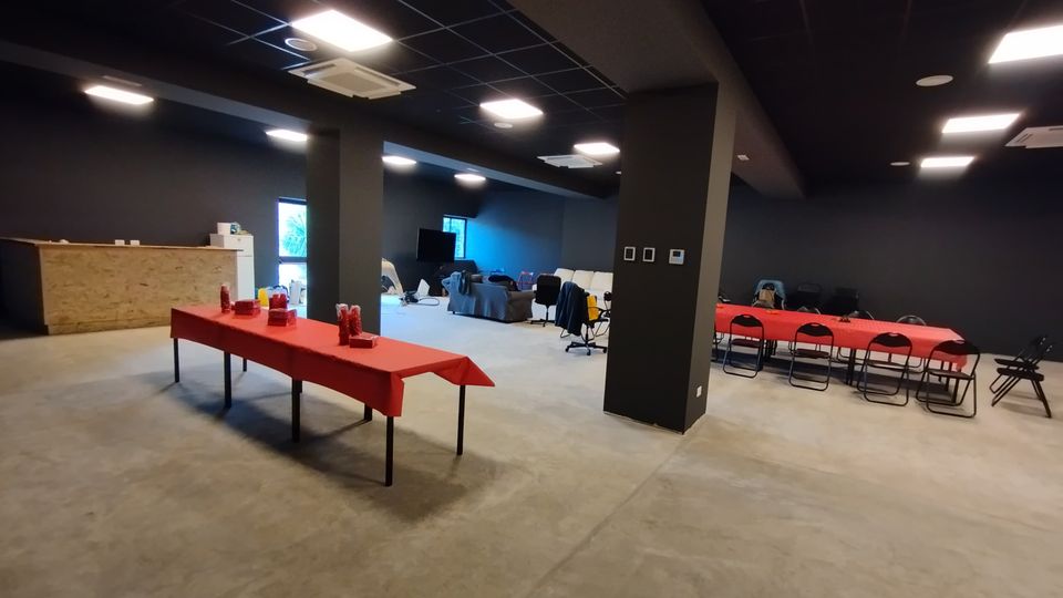 Ampio spazio interno tipo sala eventi con pareti scure e pavimento in cemento, illuminato da pannelli a soffitto e con colonne centrali. Sono presenti tavoli rettangolari coperti da tovaglie rosse con sedie, un bancone in legno su un lato e una zona lounge con divani vicino alle finestre.