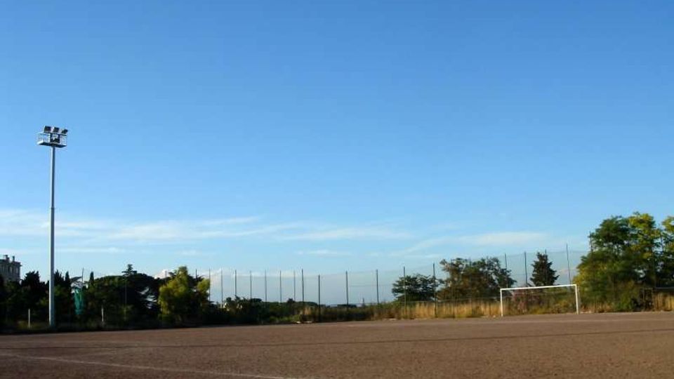 Campo sportivo all’aperto con superficie in terra battuta, una porta da calcio e pali per l’illuminazione. Sullo sfondo si vedono recinzioni alte e alberi sotto un cielo sereno.