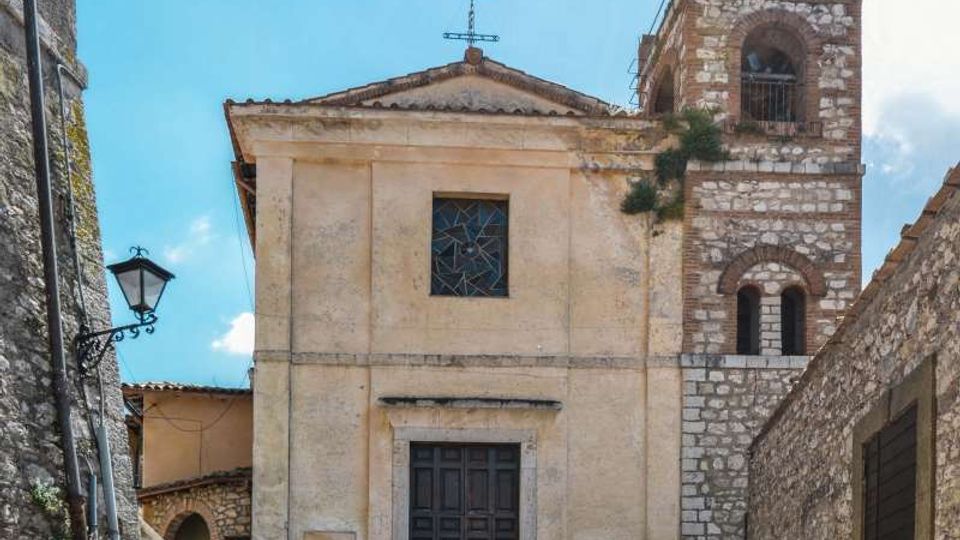 Facciata di una piccola chiesa in pietra con portale centrale, finestra quadrata e due croci sul tetto, affiancata da un campanile con aperture ad arco e cuspide con croce. Davanti c’è una piazzetta lastricata in ciottoli, circondata da muri in pietra e una lanterna a parete sotto cielo azzurro.