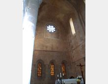 Interno di una chiesa in pietra con volta alta e pareti in muratura, illuminato da un rosone circolare e da finestre ad arco con vetri ambrati. In basso si vede l’altare con candelabri e una croce, accanto a un grande pilastro in primo piano.