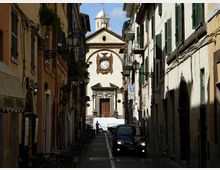Strada stretta di centro storico fiancheggiata da edifici alti con persiane verdi, balconi e lampioni a muro. In fondo alla via si vede la facciata di una chiesa con orologio centrale e campanile, con alcune auto parcheggiate lungo la carreggiata.