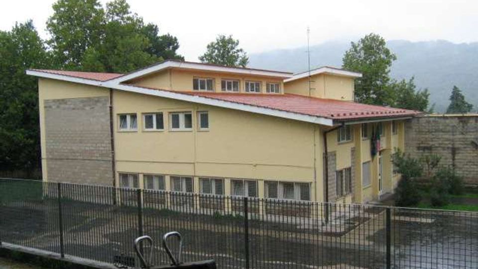 Edificio a due piani color giallo con tetto rosso a falde e file di finestre, circondato da una recinzione metallica e da un cortile asfaltato. Sullo sfondo si vedono alberi e colline o montagne parzialmente coperte da foschia.
