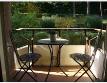 Piccolo balcone con pavimento in terracotta, ringhiera in vetro e legno, e un tavolino rotondo con vaso al centro tra due sedie pieghevoli in metallo traforato. Sullo sfondo si vede un giardino alberato con siepi e un sentiero con gradini.