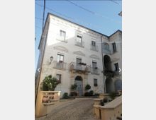 Facciata di un edificio storico intonacato di bianco su una piccola piazza acciottolata, con portale centrale ad arco, balconi con ringhiere e fioriere e finestre simmetriche. A destra si vede un passaggio ad arco che conduce a un cortile interno, con lampioni a muro e fili di illuminazione sospesi sopra.